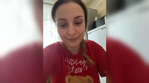 Media: Video of a smiling young woman with fair skin and light brown hair in braids, wearing a red T-shirt with \"HAPPY BIRTHDAY\" text, holding a teddy bear. Background includes a kitchen with white cabinets and a window with sheer curtains.