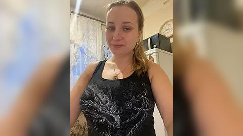 Media: Video of a fair-skinned, brown-haired woman with a slim build wearing a black tank top with a white dragon design, standing in a kitchen with a microwave and decorative clock on the wall.