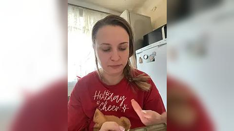 Media: Video of a young woman with light skin, brown hair, and a red sweatshirt reading \"Holiday Cheer\" holding a stuffed animal in a kitchen.