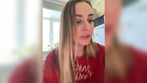 Media: A video shows a young woman with long, straight blonde hair, wearing a red t-shirt with white text. She is indoors, possibly in a kitchen, with a blurred background.