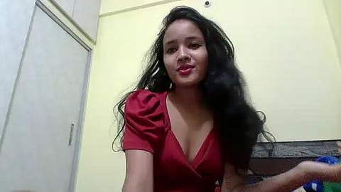 Media: Video of a smiling, light-skinned woman with long, wavy black hair, wearing a red blouse with a deep V-neck, standing in a simple room with white cabinets and a yellow wall.