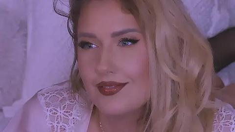 Media: Video of a fair-skinned woman with long, wavy blonde hair, wearing red lipstick and a white lace top, smiling softly.