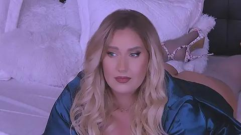 Media: Video of a blonde woman with long, wavy hair, wearing a blue satin robe, lying on a bed with white pillows and a fluffy white blanket. She has a fair complexion, full lips, and wears delicate jewelry.