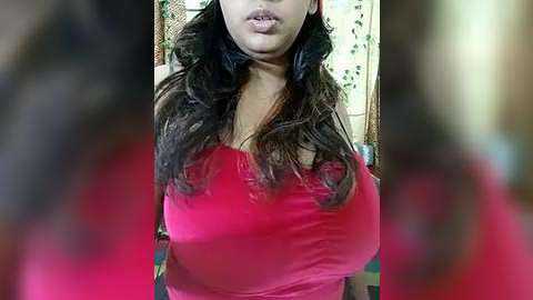 Media: A video of a woman with medium brown skin, long dark hair, and a red top, standing indoors with blurred figures in the background.