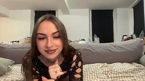 Media: Video of a young woman with long brown hair, wearing a black, polka-dot, long-sleeved top, and a nose ring, sitting on a grey couch, in a dimly lit, cluttered room with white walls and closed blinds.