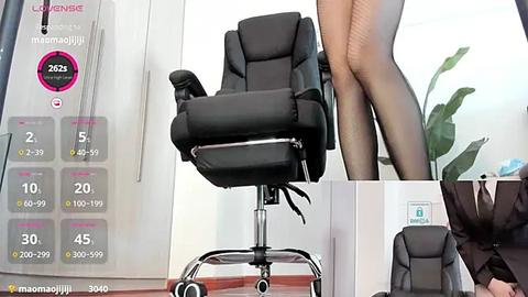 Media: Video of a woman in a black business suit standing on a black office chair. The chair features a sleek, modern design with a cushioned seat and backrest. Background includes a large potted plant and office decor.