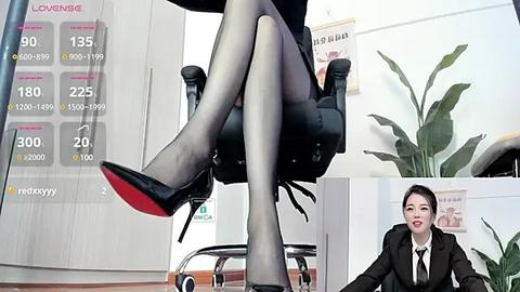 Media: Video of a woman in black business attire sitting on a black office chair, legs crossed, showing black sheer stockings and black stiletto heels. A man in a suit stands smiling in the background.