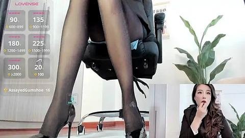 Media: Video of a woman sitting in an office chair, legs crossed in sheer black pantyhose, with a chart of pantyhose sizes on the wall.