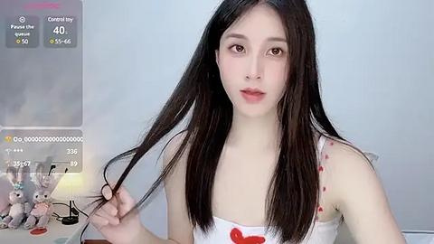 Media: Video of a slim, light-skinned Asian woman with long, straight black hair, wearing a white tank top with red accents, holding a lock of hair. Background includes a table with a lamp and a doll collection.