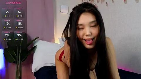 Media: A video of an Asian woman with long black hair, wearing a black top, in a playful, tongue-out pose. Background shows a bed and a plant.