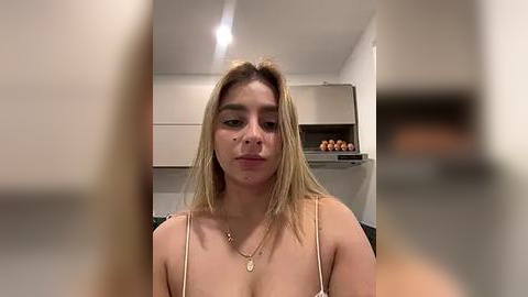 Media: Video of a young woman with straight, shoulder-length blonde hair, wearing a thin spaghetti-strap top and gold necklace. She has a light complexion and a neutral expression. Background shows a kitchen with beige cabinets and an egg carton.