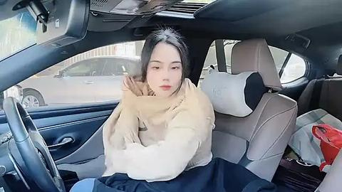 Media: Video of an Asian woman with long black hair, bundled in a beige blanket, sitting in a car's backseat. The car interior is modern with beige upholstery and visible car seats.