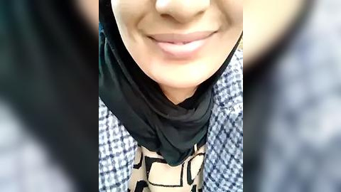 Media: Video of a smiling, light-skinned woman wearing a black headscarf and patterned blouse. The background features a checkered blue and white fabric.