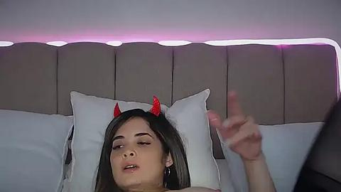 Media: Video of a young woman with light skin, long dark hair, and red devil horns, lying on a bed with a gray padded headboard. She has a relaxed expression and one hand raised.