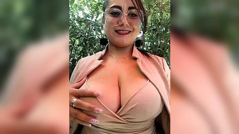 Media: Video of a smiling, voluptuous woman with large breasts, wearing a pink blouse, revealing cleavage, and clear glasses. Background features green foliage, suggesting an outdoor setting.