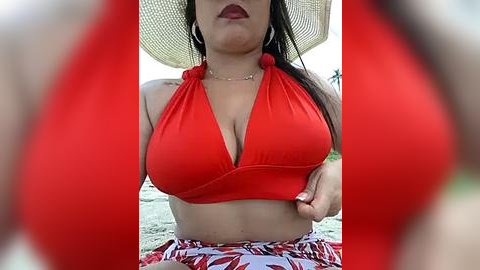 Media: Video of a woman in a red halter bikini top with large breasts, sitting outdoors. She wears a straw hat and has medium brown skin.