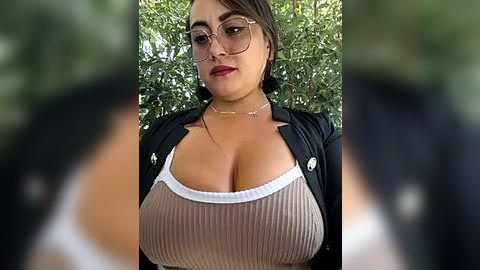 Media: Video of a young woman with light skin, brown hair, and glasses, wearing a low-cut ribbed beige top revealing ample cleavage. She has a confident expression and is outdoors with a blurred green background.