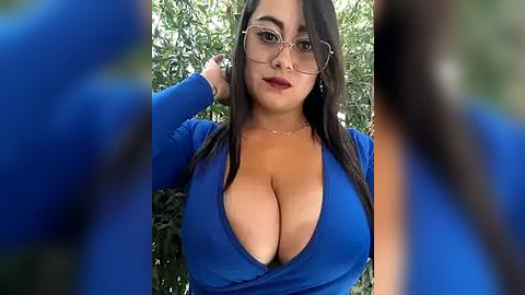 Media: A video of a Latina woman with long brown hair, wearing large glasses and a low-cut blue top, posing outdoors with blurred trees in the background.