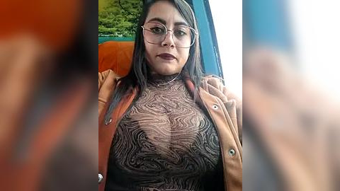 Media: Video of a woman with fair skin, medium build, wearing glasses, a brown jacket, and a sheer, patterned top, sitting in a car with orange seat.