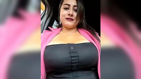 Media: Video of a Latina woman with medium skin tone, long black hair, wearing a pink cardigan over a black ribbed tank top, driving in a car with a blurred, pink background.