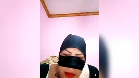 Media: Video of a person wearing a black hooded mask, red lipstick, and a white shirt, against a pink wall with a wooden ceiling.