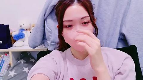 Media: Video of a young East Asian woman with long brown hair, wearing a light pink T-shirt, covering her mouth with her hand, in a cozy bedroom with plush toys and a blue blanket in the background.