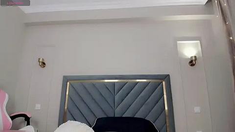 Media: Video of a modern bedroom featuring a gray, tufted headboard with gold trim, two wall-mounted sconces, and a plush, dark navy chair.