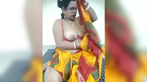 Media: A video of a South Asian woman in a yellow sari, revealing her large breasts, sitting on a checkered floor, wearing glasses, indoors.