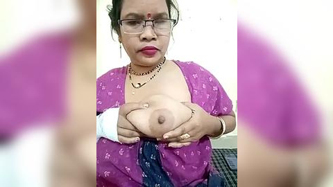 Media: Video of an Asian woman with glasses, red dot on forehead, wearing purple blouse, holding her large breasts, in blurred background.