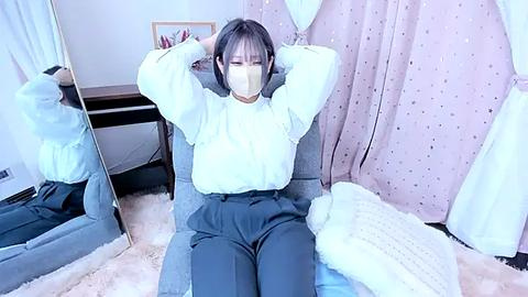 Media: Video of an Asian woman with short black hair, wearing a white mask and a loose white blouse, sitting on a gray chair with arms raised, surrounded by light pink curtains and fluffy white blankets.