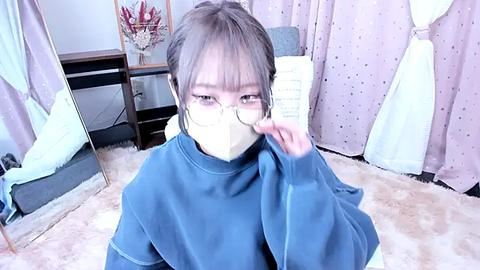 Media: A video of an Asian woman with glasses and a mask, sitting on a plush carpet, wearing a blue sweatshirt, holding a book in a cozy, pastel-pink room with a large mirror and a flower arrangement.