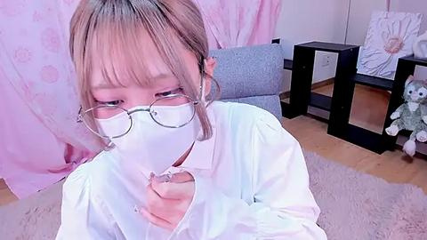 Media: Video of a young Asian woman in a white shirt, wearing glasses, with a mask covering her mouth, sitting in a pastel room with a plush toy, a black table, and a pink curtain.