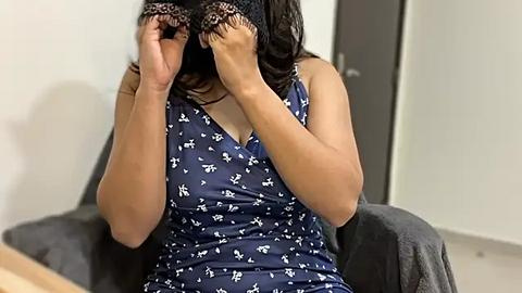 Media: Video of a South Asian woman with medium skin tone, wearing a dark blue sleeveless dress with white floral patterns, covering her eyes with a black lace mask, sitting on a gray chair in a minimalist office setting.