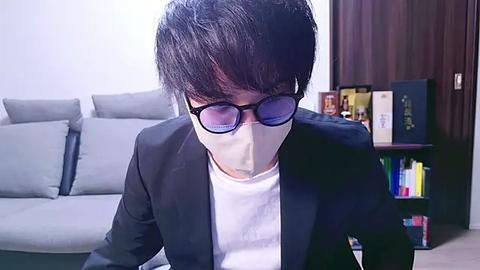 Media: Video of an Asian man with short black hair, wearing dark glasses, a black suit, and a white mask, sitting indoors, surrounded by light gray couches and bookshelves.