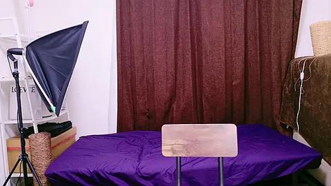 Media: Video of a small, simple room with a purple bed and a wooden table, dark brown curtains, a white wall, and a white shelf with a blue light stand and a basket.