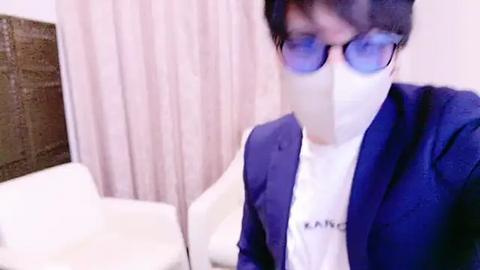 Media: Video of an Asian man in a blue suit and white mask, wearing blue-tinted glasses, standing in a modern, minimalist room with white furniture and light-colored curtains.