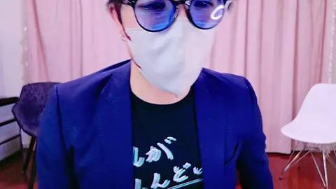 Media: A video of an Asian woman with short dark hair, wearing blue-tinted glasses, a blue blazer, and a black T-shirt with Japanese text. She has a white mask covering her nose and mouth. The background features pale pink curtains and a white chair.