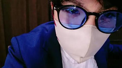 Media: Video of a person wearing large blue-tinted sunglasses, white face mask, and a blue blazer, against a dark brown wooden background.