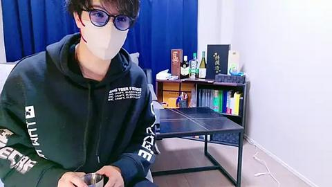 Media: Video of a person wearing a face mask, black hoodie, and glasses, seated at a table in a room with blue curtains, a bookshelf, and assorted items.