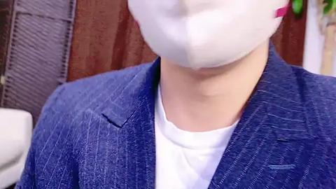 Media: Video of a person wearing a white surgical mask, white t-shirt, and blue pinstriped blazer, with blurred background showing a white couch and brown wooden wall.