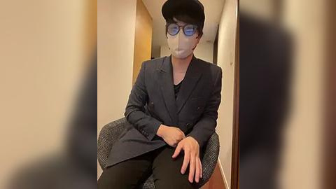 Media: Video of a person wearing a black face mask, black suit, and glasses, seated in a chair with a blurred background.