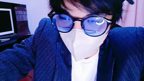 Media: Video of an Asian man with short black hair, wearing blue pinstriped suit jacket, white shirt, and blue-tinted glasses. He is wearing a beige surgical mask, and the background features a TV and a window with white curtains.