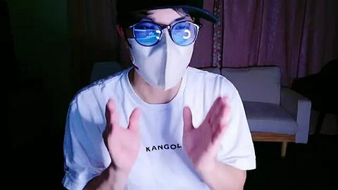 Media: Video of a person wearing a white mask, blue glasses, and a white T-shirt with \"KANGOL\" printed on it, hands raised in the air, indoors with a white sofa and pink curtains in the background.