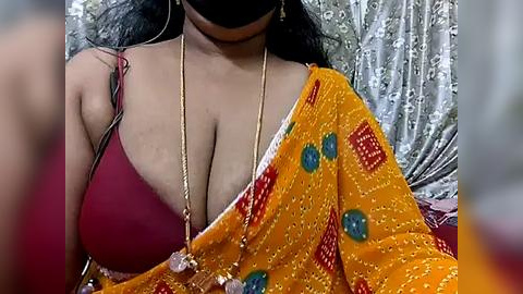 Media: Video of a South Asian woman with medium skin tone, wearing a red bra and a vibrant orange saree with green and red patterns, against a silver sequined background.