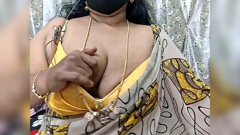 Media: Video of a South Asian woman with medium skin tone, wearing a yellow bra, black mask, and a yellow and white floral-patterned sari.