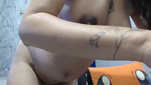 Media: Video of a nude woman with medium-sized breasts, light skin, and dark pubic hair. She has tattoos on her arms and is holding a pillow with an orange cat design. Background includes a textured wall and a white bed.