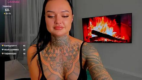 Media: Video of a tattooed woman with long black hair, wearing a black top, standing in front of a lit fireplace, with viewers' comments displayed on screen.