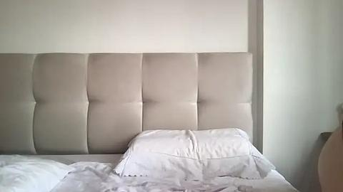 Media: Video of a minimalist bedroom with a beige tufted headboard, white bedding, and a neatly arranged pillow. The background features a plain white wall and a partially visible beige nightstand.