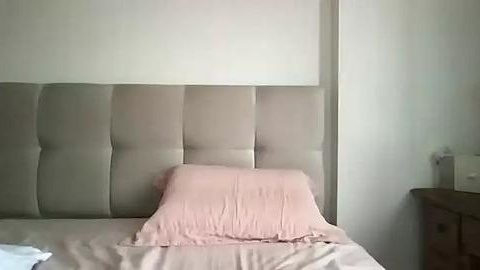 Media: Video of a minimalist bedroom featuring a beige tufted headboard, a pink pillow, and a matching pink bedspread, with a white wall and a dark wooden nightstand on the right.