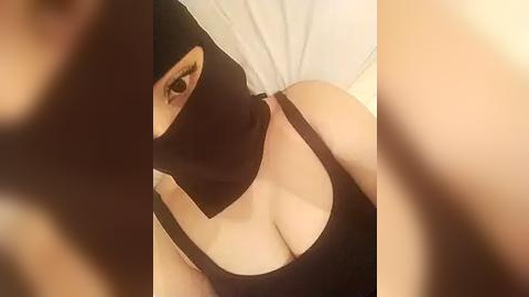 Media: Video of a woman in a black face mask, black top, with her large breasts partially visible, taken from an angle, indoors with a blurred background.